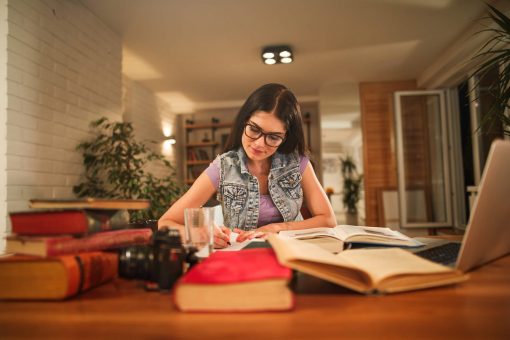 como focar nos estudos: mulher jovem sentada em mesa de casa em volta de vários livros